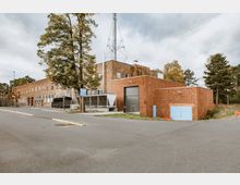 Backsteingebäude in einem Gewerbe- oder Industriegebiet mit flachem Dach, großem Tor und mehreren Fenstern. Im Vordergrund ein Parkplatz und ein Gehweg, umgeben von Bäumen und Grünflächen.