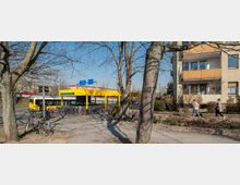 Gelbe Bushaltestelle mit Wartehäuschen und Fahrrädern davor, umgeben von Bäumen und Fußwegen; im Hintergrund ein mehrstöckiges Wohngebäude mit Balkonen.