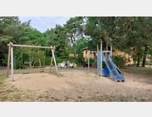 Ein Spielplatz mit einer Schaukel, einer blauen Rutsche und Kletterelementen, umgeben von Sandboden und Wiese. Im Hintergrund sind Bäume, Büsche, Holztierfiguren und ein gelbes Gebäude zu sehen.