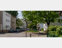 Wohnstraße mit Mehrfamilienhäusern, parkenden Autos und Bäumen an den Gehwegen. Ein Straßenschild mit der Aufschrift „Hechelstraße“ steht im Vordergrund, umliegende Begrünung sorgt für eine ruhige Atmosphäre.