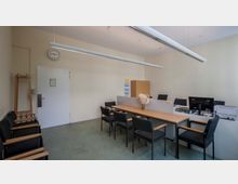 Büro- oder Besprechungsraum mit schlichtem Design, bestehend aus einem langen Tisch mit schwarzen Stühlen, Computern auf einem Schreibtischbereich und einem Aktenschrank im Hintergrund. An der Wand befindet sich eine Uhr, und der Raum hat helle Wände und Hängelampen.