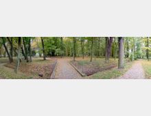 Panorama eines Parks mit hohen, dicht stehenden Bäumen, herbstlich gefärbtem Laub auf dem Boden und mehreren geschwungenen Wegen. In der Mitte befindet sich eine schmale Brücke mit Geländern, die einen kleinen Graben überspannt.