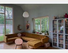 Wohnzimmer mit einer senfgelben Eckcouch, einem runden Holztisch und großen Fenstern, die den Blick ins Grüne ermöglichen. Rechts steht ein Bücherregal mit Glastüren, auf der Fensterbank befinden sich Pflanzen und Dekorationsgegenstände.