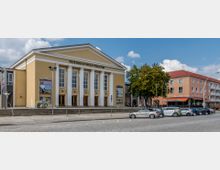 Das Bild zeigt das Friedrich-Wolf-Theater, ein gelbes Gebäude mit klassizistischer Architektur, gekennzeichnet durch sechs weiße Säulen und eine dreieckige Frontfassade. Neben dem Gebäude befinden sich ein Baum, geparkte Autos sowie ein benachbartes Gebäude mit einem Café im Erdgeschoss.