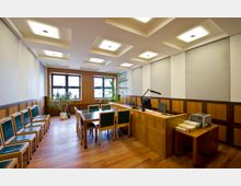 Moderner Gerichtssaal mit Holzvertäfelung, einem Richterpult mit Computerarbeitsplatz und Mikrofon, einem Besprechungstisch mit grünen Stühlen und hellem Holzfußboden. Große Fenster und Zimmerpflanzen sorgen für ein lichtdurchflutetes Ambiente.