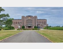 Backsteingebäude im Stil der frühen Moderne mit symmetrischer Fassade, großen Fenstern und einem zentralen Eingangsbereich mit Überdachung; davor eine gepflasterte Auffahrt, umgeben von gepflegten Grünflächen und Bäumen.