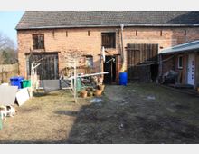 Hinterhof eines alten Gebäudes mit einer Fassade aus rotem Backstein und einem grauen Ziegeldach. Im Hof stehen Gartenutensilien, Töpfe, ein Wäschegestell und eine blaue Regentonne, während links Mülltonnen zu sehen sind; ein Hund befindet sich im Vordergrund.