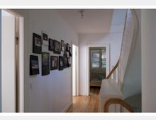 Flur in einem Wohnhaus mit Holzboden und weiß gestrichenen Wänden, links eine Wand mit gerahmten Bildern und Kalendern, rechts eine Treppe mit weißem Geländer und Holzhandlauf. Im Hintergrund befindet sich ein offenes Zimmer mit Bett und einem Fenster mit Lamellenrollo.