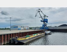 Industriehafen mit einem großen blauen Hafenkran auf einer Betonpier, einem gelben Lastkahn und einem Schlepper im Wasser. Im Hintergrund sind ein weißes Lagergebäude, ein Schüttguthaufen und eine Abgrenzungsmauer zu sehen.