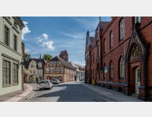 Straßenszene in einer Altstadt mit gepflasterter Straße, links Fachwerkhäuser und rechts ein Gebäude aus rotem Backstein mit Spitzbögen. Im Hintergrund ist der Turm einer Kirche zu sehen, und geparkte Autos befinden sich entlang der Straße.