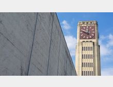 Hochformat eines Gebäudes mit einer großen Wanduhr auf einem Turm. Die rechteckige Uhr hat ein rotes Zifferblatt und weiße Zeiger; daneben ein hoher, beiger Betonturm mit mehreren Fensterreihen. Links des Turms befindet sich eine schlichte, beige Wand.