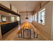 Großzügiger, heller Raum mit moderner Küche und langem Holztisch mit Sitzbänken. Links befindet sich eine Küchenzeile mit dunklen Schränken und Holzarbeitsplatte, rechts eine Reihe von Fenstern mit weißen Rahmen und dekorierten Fensterbänken.