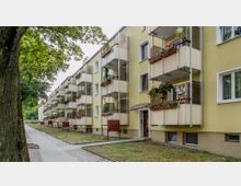 Mehrstöckiges Wohngebäude mit gelber Fassade und Reihen von Balkonen, die mit Pflanzen geschmückt sind. Vor dem Gebäude verläuft ein gepflasterter Gehweg, flankiert von Rasenflächen und Bäumen.