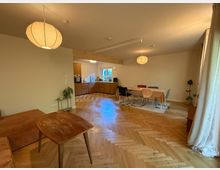 Ein offener Wohn- und Essbereich mit Holzfußboden in Fischgrätmuster, einer zentralen Esstischgruppe mit verschiedenen Stühlen und einer modernen Küche im Hintergrund. Zwei runde, hängende Lampen und bodenlange Vorhänge an den Fenstern verleihen dem Raum eine helle Atmosphäre.