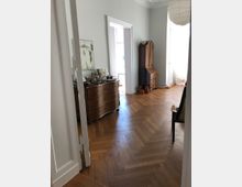 Ein helles Zimmer mit Parkettboden in Fischgrätenmuster, verziert mit hellgrauen Wänden. Zu sehen sind ein brauner, geschwungener Kommodenschrank, eine antike Holzsekretärkommode und ein weißer Lampenschirm an der Decke. Im Hintergrund führen geöffnete Türen zu einem weiteren, lichtdurchfluteten Raum.