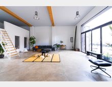 Großer, moderner Wohnraum mit hohen Decken, sichtbaren Holzbalken und einer Betonbodenfläche. Der Raum umfasst eine schwarze Ledercouch, eine minimalistische Holztreppe, helle tatamiartige Teppiche und große Fensterflächen, die viel natürliches Licht hereinlassen; Pflanzen und schlichte Möbel ergänzen das minimalistische Design.