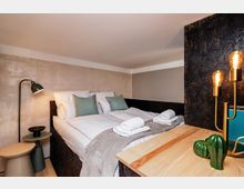 Modernes, kompaktes Schlafzimmer mit Doppelbett unter einer niedrigen Dachschräge, in neutralen Wandfarben und mit weißen Bettwaren, grauer Decke und zusammengerollten Handtüchern. Neben dem Bett stehen eine Stehlampe und kleine Beistelltische; im Vordergrund sind eine Holzablage und eine schwarze Trennwand mit warm leuchtenden Edison-Lampen zu sehen.