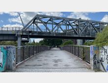 Fußgängerbrücke mit Geländern, die zu einer unter einer Stahlkonstruktion verlaufenden Bahnbrücke führt. Die Umgebung zeigt Bäume, Teile eines Gebäudes und Wände mit Graffiti.