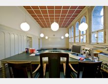 Heller Besprechungsraum mit holzverkleideten Wänden, hohen Fensterbögen, einer Rasterdecke in Rot-Weiß und mehreren runden Hängelampen. In der Mitte steht ein großer Konferenztisch mit Büchern, Monitoren und Büromaterial, umgeben von Holzstühlen.