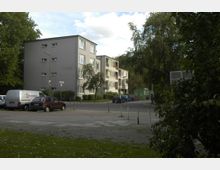 Wohnsiedlung mit einem grauen, mehrstöckigen Wohngebäude, umgeben von Bäumen und einer Straße mit parkenden Autos. Im Vordergrund ein Fußweg und eine Rasenfläche, im Hintergrund ein grüner Container.