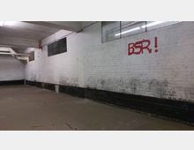 Ein leerer, betonierter Kellerraum mit weißen Ziegelwänden, an denen Schwarzspuren zu sehen sind. Oben befindet sich ein rotes Graffiti mit der Aufschrift "BSR!", und entlang der Decke verlaufen Rohre und Kabel.