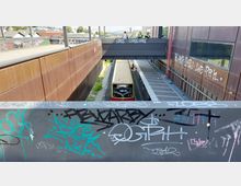 Blick auf einen S-Bahnhof mit einem einfahrenden Zug, umgeben von modernen Gebäuden aus rostbraunem Metall. Eine Fußgängerbrücke mit Graffiti überspannt die Gleise, und das Geländer im Vordergrund ist ebenfalls mit Graffiti bedeckt.