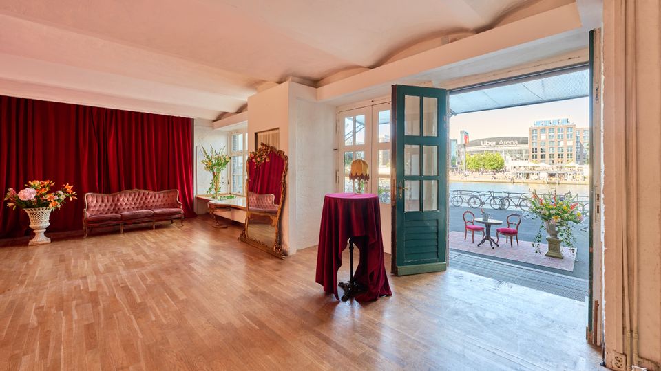 Großer, heller Innenraum mit Holzparkett, hohen weißen Decken und burgunderroten Vorhängen, ausgestattet mit einem roten Sofa, einem hohen Spiegel und dekorativen Blumenarrangements. Eine offene türkisfarbene Glastür führt zu einer Terrasse am Wasser mit Café-Tischen; im Hintergrund sind moderne Gebäude zu sehen.