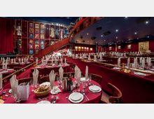 Ein festlich eingerichteter Theatersaal mit rotgepolsterten Sitzbereichen und rot gedeckten Tischen, dekoriert mit weißen Stoffservietten und Weingläsern. Im Hintergrund befinden sich eine Bühne mit rotem Vorhang und eine Galerie mit beleuchteten Bühnenkostümen und Postern an den Wänden.