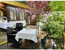 Außenbereich eines kleinen Restaurants oder Cafés mit einem Tisch und Stuhl auf einer gepflasterten Terrasse. Im Hintergrund sind ein Gebäude mit großen Fenstern, grüne Pflanzen und ein Baum mit roten Blättern zu sehen, daneben eine kleine weiße Ente auf dem Boden.