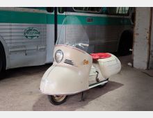 Innenraum einer alten Garage oder eines Depots mit Betonboden und gemauertem Pfeiler, in dem ein cremefarbener Vintage-Roller mit Windschutzscheibe und rotem Sitz steht. Im Hintergrund ist die Seitenwand eines grün-silbernen Busses mit Nieten und Fenstern zu sehen.
