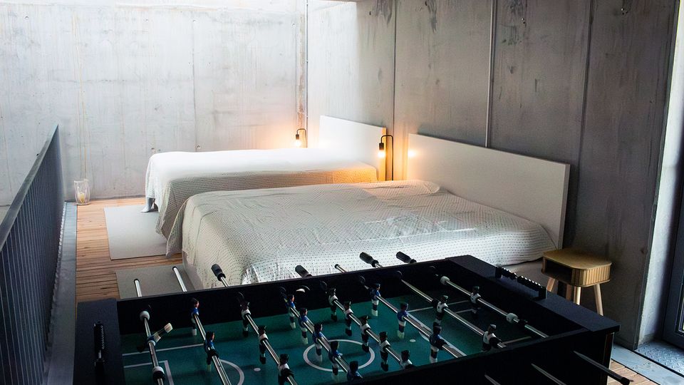 Minimalistisches Schlafzimmer mit Sichtbetonwänden und Holzfußboden, zwei großen Betten mit weißen Decken und schlichten Kopfteilen sowie warmen Wandlampen. Im Vordergrund steht ein Tischkicker, daneben ein kleiner Hocker nahe einem Fenster.