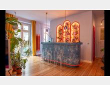 Moderne Bar im Wohnbereich mit einer blau gemusterten Theke und leuchtenden, orange umrahmten Regalen voller Flaschen. Holzfußboden, große Fenster mit gelben Vorhängen und zahlreiche Pflanzen verleihen dem Raum eine helle und lebendige Atmosphäre.