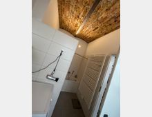 Badezimmer mit weiß gefliesten Wänden, einer Badewanne, einem wandmontierten Handtuchheizkörper und einem Föhn an der Wand. Die Decke besteht aus sichtbaren Ziegelsteinen mit einer Rundbogenstruktur.