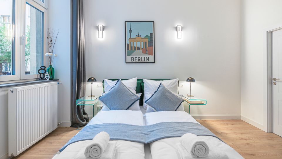 Helles, modern eingerichtetes Schlafzimmer mit zwei zusammengestellten Einzelbetten, grauen Kissen und gerollten Handtüchern am Fußende. Neben großen Fenstern mit Vorhang stehen Glas-Nachttische mit Lampen; an der Wand hängen ein Berlin-Poster und zwei Wandleuchten.