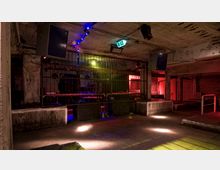 Industrieähnlicher Clubraum in einem Keller mit Betonwänden und -säulen, dunkler Beleuchtung in Rot, Gelb und Blau sowie einer Bühne hinter einem Metallgitter. Oben sind Lautsprecher befestigt, und ein Notausgangsschild leuchtet über der Szene.