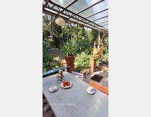 Überdachte Gartenterrasse mit transparentem Wellplatten-Dach und sichtbaren Stahlträgern, davor ein Tisch mit Geschirr und einer Schale Erdbeeren. Im Hintergrund ein verwilderter Garten mit Bäumen und Sträuchern, ein Hängesessel sowie mehrere Pflanzgefäße und niedrige Ziegelpfeiler.