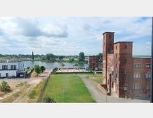 Alte Industriegebäude aus rotem Backstein mit Türmen in einer parkähnlichen Umgebung am Ufer eines Flusses, im Hintergrund eine Brücke und grüne Landschaft. Links im Bild moderne, weiße Gebäude und ein Sandbereich mit Sitzmöglichkeiten.