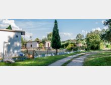 Ein Weg führt an einer Reihe von Kleingärten entlang, die von Metall- und Maschendrahtzäunen umgeben sind. Im Vordergrund ist ein kleines Gartenhaus mit der Nummer „26“ sichtbar, umgeben von Bäumen, Sträuchern und Blumen.