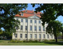 Barockgebäude mit hellem Putz, symmetrischen Sprossenfenstern und einem roten Ziegeldach, umgeben von einer Grünanlage mit Bäumen und Rasenflächen.