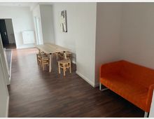 Ein schlichter Raum mit Holzboden, einem hellen Holztisch mit kleinen Holzstühlen und einem orangefarbenen Sofa in der rechten Ecke. Im Hintergrund ist ein Heizkörper an der weißen Wand und eine halb geöffnete Tür sichtbar.