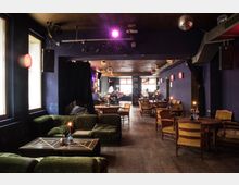 Gemütliche Bar mit abgedunkelter Beleuchtung, dunkelblauen Wänden und Holzboden. Die Einrichtung besteht aus grünen Sofas, hölzernen Tischen, gelben Stühlen und Kerzen auf den Tischen, mit dekorativen Lampen und einer Discokugel an der Decke.