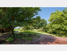 Ein parkähnlicher Bereich mit einem geschwungenen, gepflasterten Weg, umgeben von großen Bäumen und grüner Vegetation. Im Hintergrund ist eine offene Wiese bei klarem, sonnigem Wetter zu sehen.