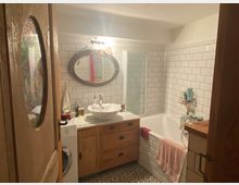 Badezimmer mit weiß gefliesten Wänden und dekorativem Bodenmuster, ausgestattet mit einer freistehenden Holzkommode, einem ovalen Spiegel, einer Badewanne und einer Waschmaschine.