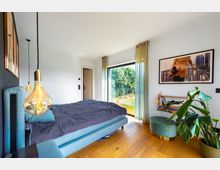 Modernes Schlafzimmer mit einem blauen Polsterbett, hölzernem Boden und einem großen Fenster, das auf eine grüne Landschaft blickt. Im Raum befinden sich ein hängendes Glühbirnenlicht, ein großer Pflanzenkübel, ein Bücherregal und ein modernes Bild an der Wand.