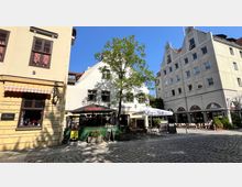 Straßenszene in einer Altstadt mit gepflastertem Platz, flankiert von historischen Gebäuden mit hellen Fassaden. Im Vordergrund befinden sich ein Baum, Restauranttische, Sonnenschirme und ein kleiner Zaun, während Geschäftseingänge und Cafés im Hintergrund sichtbar sind.