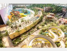 Innenansicht eines tropischen Freizeitparks unter einer großen Halle, mit einer langen gelben und silbernen Wasserrutsche, mehreren Pools, Liegestühlen und Palmen. Die Umgebung ist mit tropischen Pflanzen, bunten Gebäuden und kleinen Wegen gestaltet.