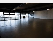 Großer, leerer Innenraum in einer Industriehalle mit dunkler Decke, Betonboden und langen Fensterfronten entlang einer Seite. Metallgeländer stehen vor den Fenstern; an der Stirnseite sind eine breite weiße Wand, eine Treppe und Deckenleuchten zu sehen.