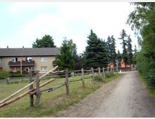 Schotterweg mit Holzzaun entlang einer Wiese, im Hintergrund mehrstöckige Wohngebäude mit Balkonen und grünen Bäumen. Rechts des Weges dichter Bewuchs, links einige hohe Tannen.