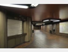 U-Bahn-Passage mit braunen Fliesen, Wänden aus Ziegelsteinen und abgehängten Decken mit integrierter Beleuchtung. Im Vordergrund ein geschlossenes Rolltor mit Graffiti, daneben ein Wandtelefon und ein Stadtplan.
