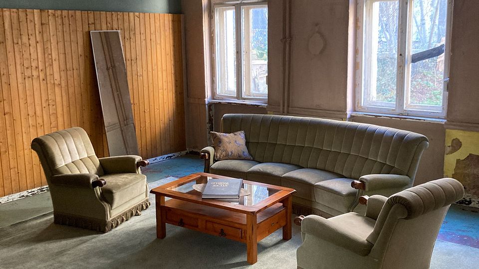 Ein spärlich eingerichteter Raum mit hohen, alten Doppelfenstern, teils abgenutzten Wänden und Holzvertäfelung an einer Seite. In der Mitte steht eine Sitzgruppe aus Sofa und zwei Sesseln auf einem großen Teppich um einen niedrigen Holztisch.